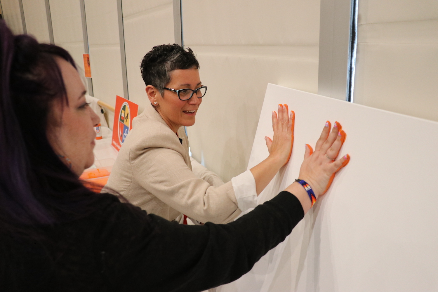 Hali Topinka and Sheena Yew place an orange hand print on a canvas.